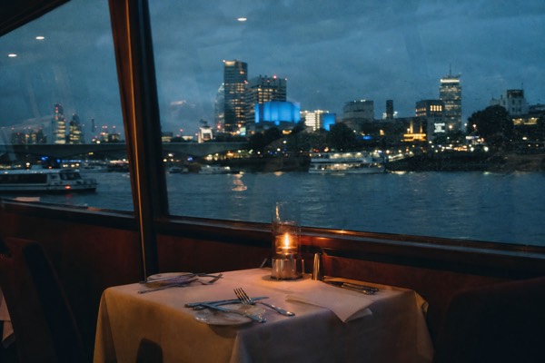 Dinner table set by a window on a Thames cruise boat with the lit riverfront behind