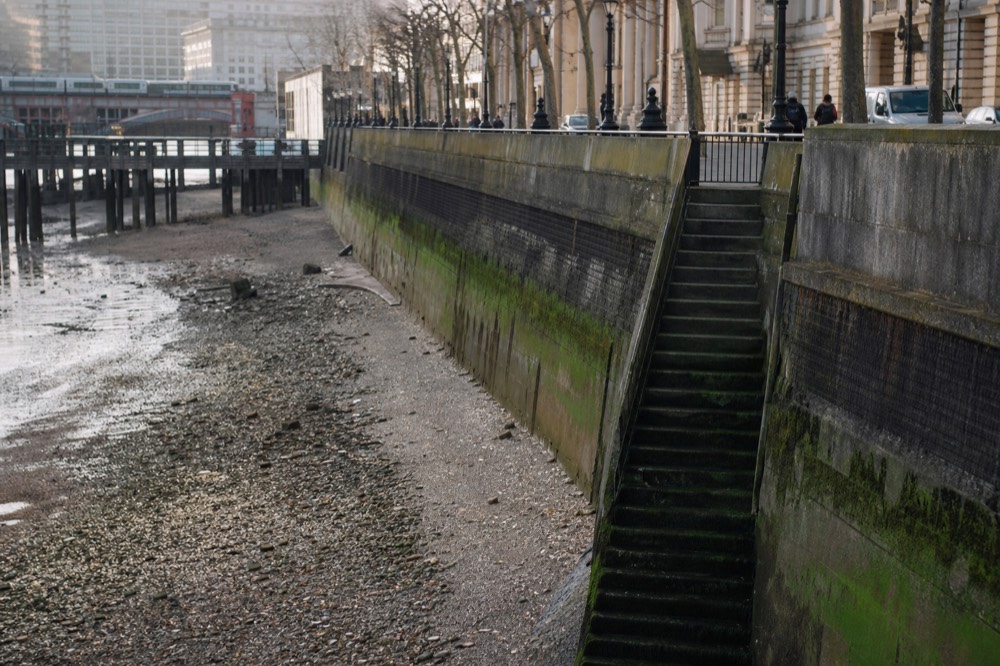 Steep algae-covered stone river stairs descending to exposed gravel foreshore at low tide, a high sheer embankment wall above
