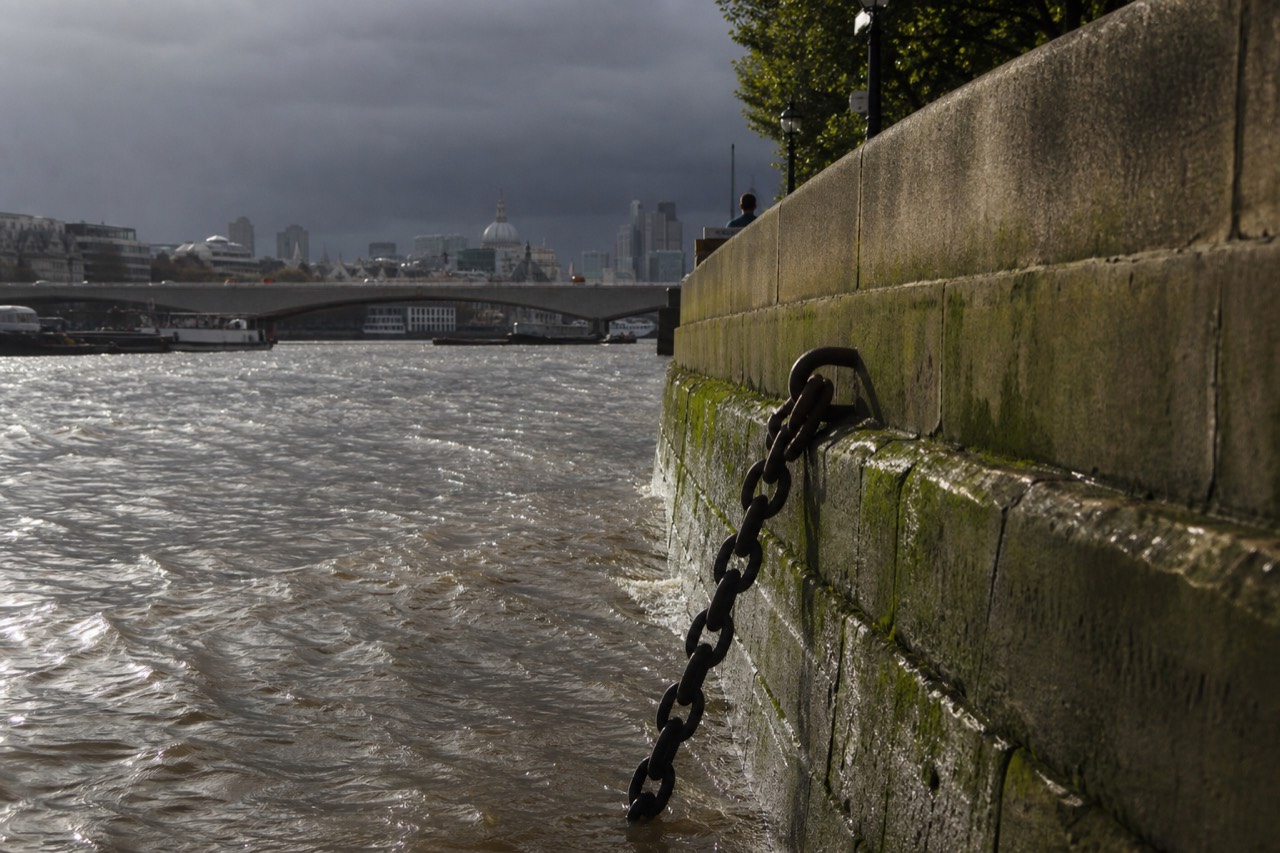 Fast brown tidal water running past a weathered stone river wall in central London under flat grey cloud, no swimmers, a mooring chain half submerged