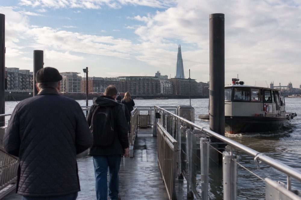 Passengers waiting on a floating river pontoon in morning light with a gangway ahead and a boat coming alongside