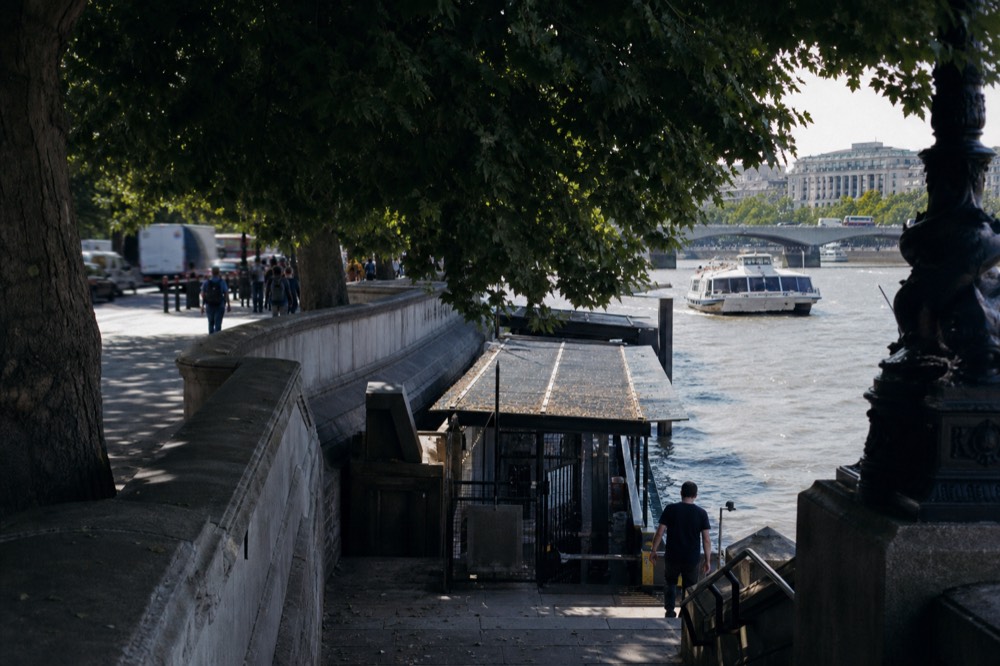 Stone steps and railings leading down to a river pier entrance on a busy afternoon, the embankment wall curving away behind