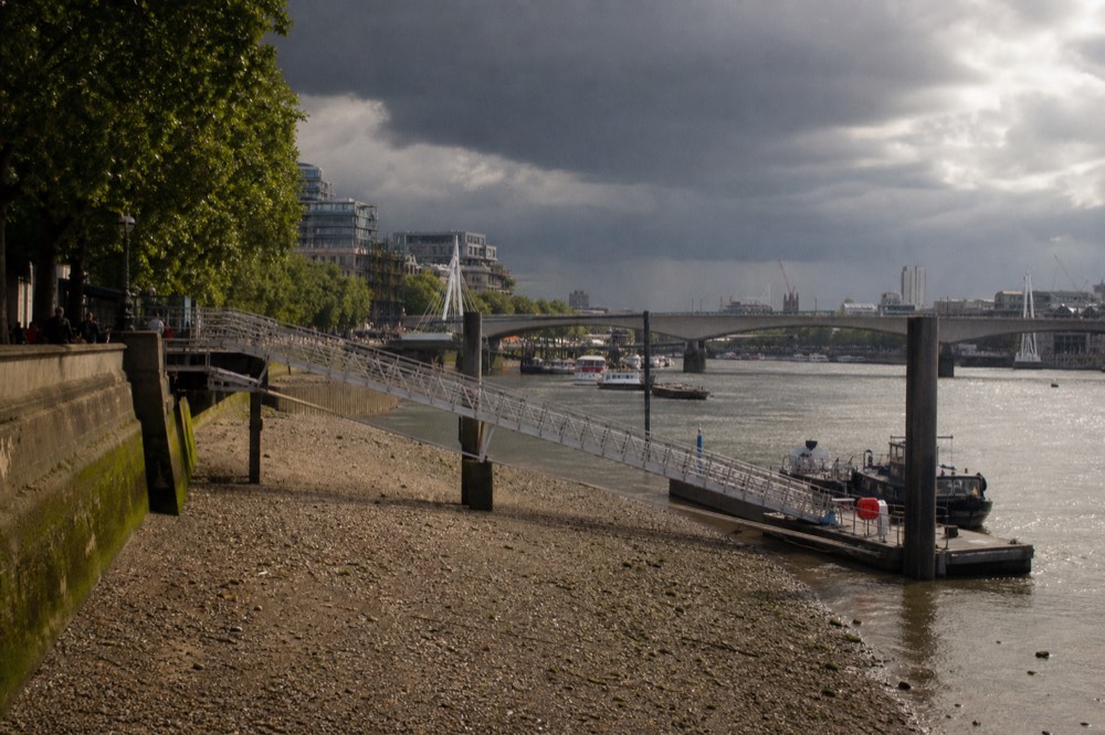 Exposed gravel foreshore at low tide below an embankment wall, a steep gangway running down to a pontoon under grey sky