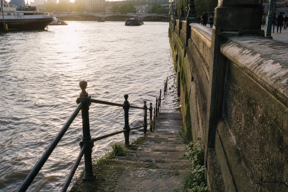 Worn Victorian stone river stairs descending into brown tidal water beside a granite embankment wall, iron handrail and green weed on the lower treads