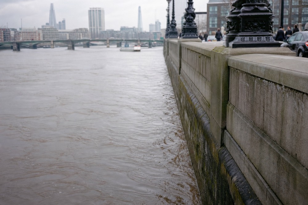 A sheer granite embankment wall at high water on a grey day, brown current running fast along the stonework with no steps or ladder in view