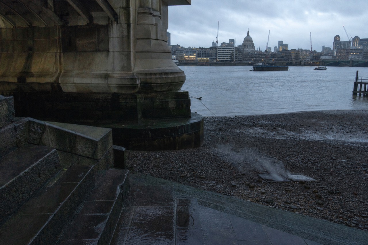 The tidal Thames at low water beneath a Victorian bridge abutment, wet stone steps running down to a shingle foreshore under grey cloud