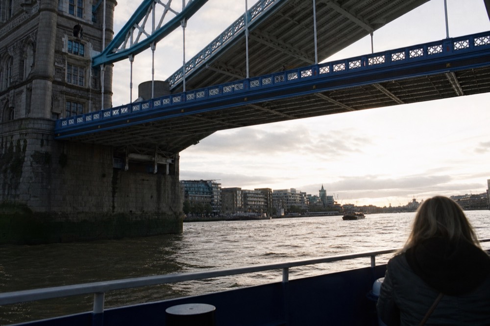 View from a boat deck looking up at the underside of Tower Bridge, blue ironwork and high walkways against pale sky