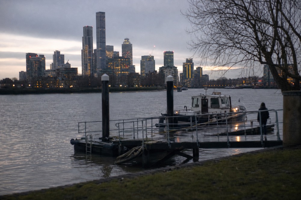 A quiet floating pier on a grey winter afternoon, wet decking and a single moored boat, bare trees on the embankment behind