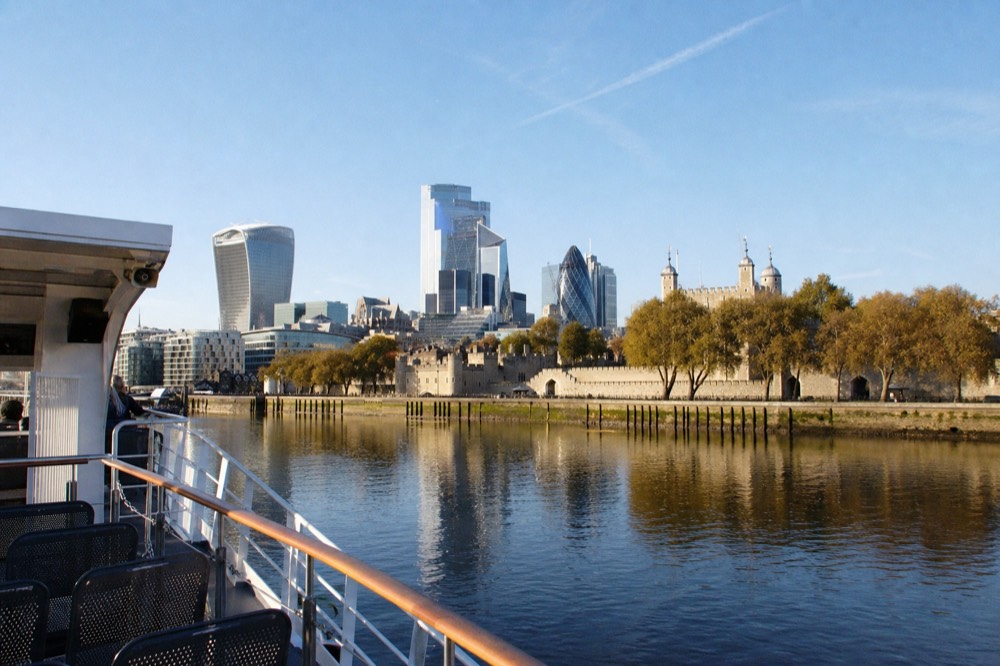 The lit south bank reflected in flat dark water on a clear evening, a boat rail and empty deck seats in the foreground