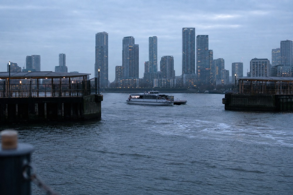 A small modern passenger ferry crossing between two docklands piers on flat water with tall glass towers rising on the far bank