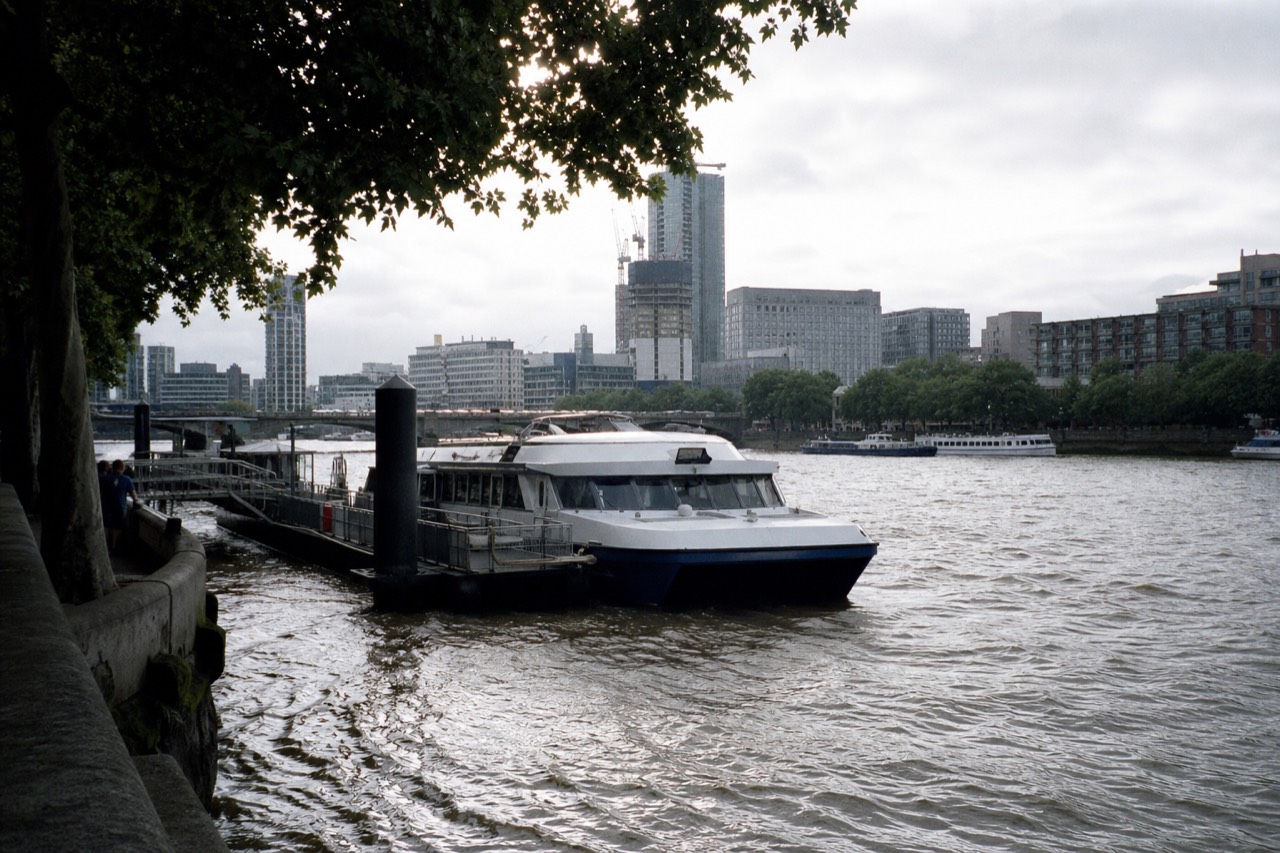 A fast twin-hulled passenger catamaran alongside a floating pier on the river, city buildings behind under flat morning cloud