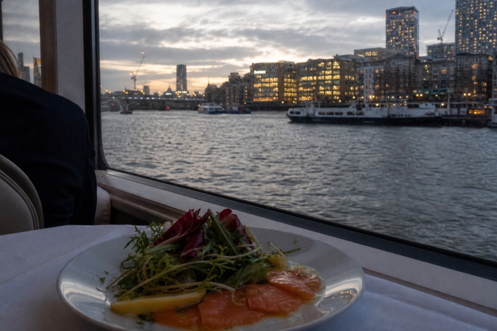 A cured salmon starter plated with dressed leaves on a white cloth beside a boat window, dark water outside