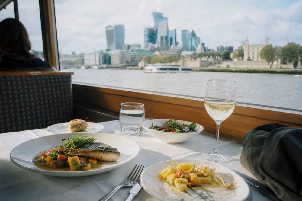 A two-course lunch laid on a boat table in bright daylight, the Thames and riverside buildings visible through a wide window