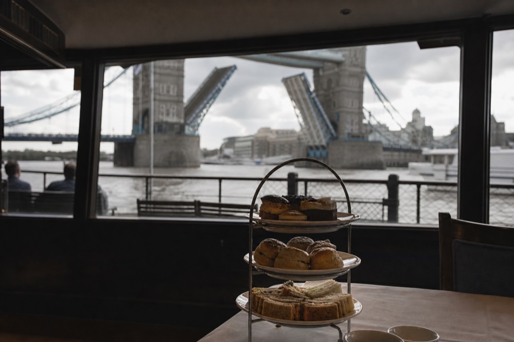 A three-tier stand of finger sandwiches, scones and small cakes on a boat table with a raised bridge tower beyond the window
