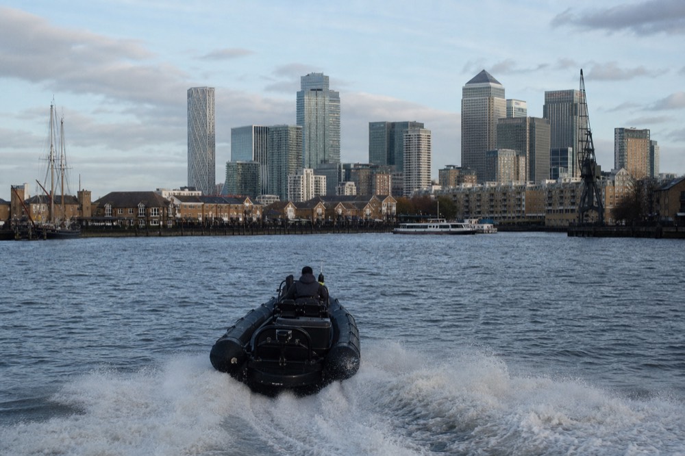 A small rigid inflatable boat running fast across open river water on a bright day, wake spreading wide, docklands towers in the distance