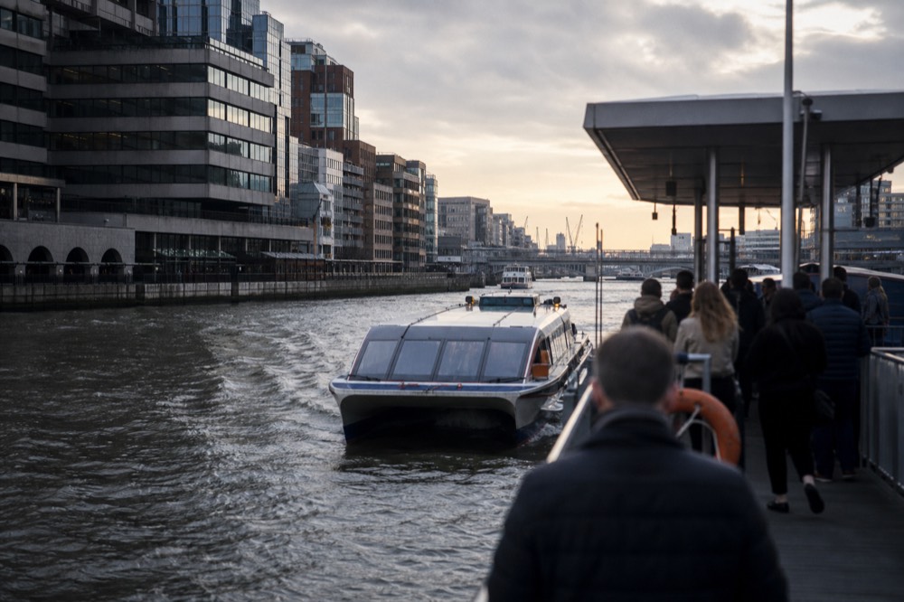 A fast catamaran river bus drawing alongside a floating pier on a grey morning, wake churning, commuters waiting on the pontoon above