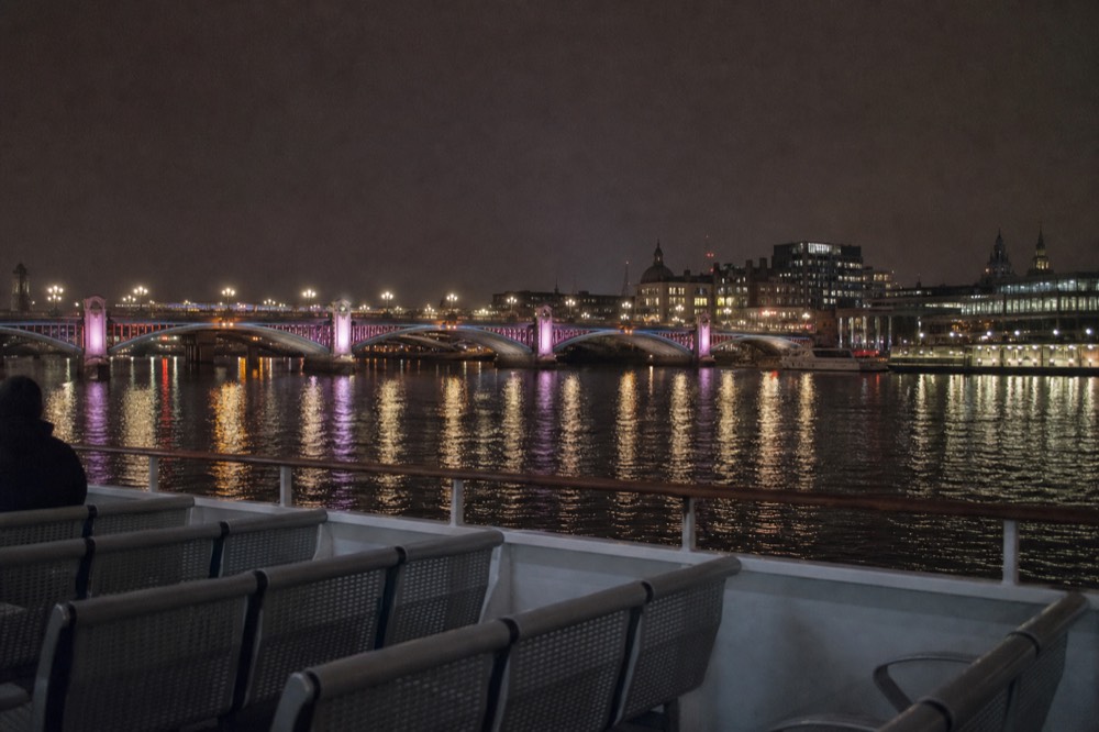 A floodlit river bridge reflected in flat black water on a clear night, boat rail and empty deck seats in the foreground
