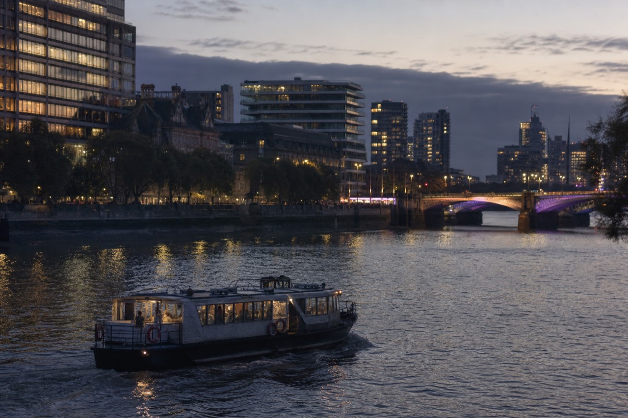 A river boat crossing flat water at dusk with the lit south bank behind it and a floodlit bridge span in the distance