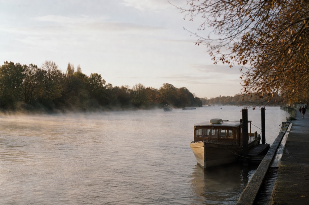A straight wide reach of river between wooded banks in early morning light, a wooden launch at a mooring post and mist lifting off flat water