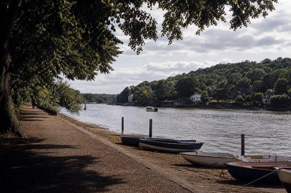 A wide bend of the tidal river seen from a towpath under mature trees, a wooded hill rising on the far bank and small rowing boats drawn up on a slipway