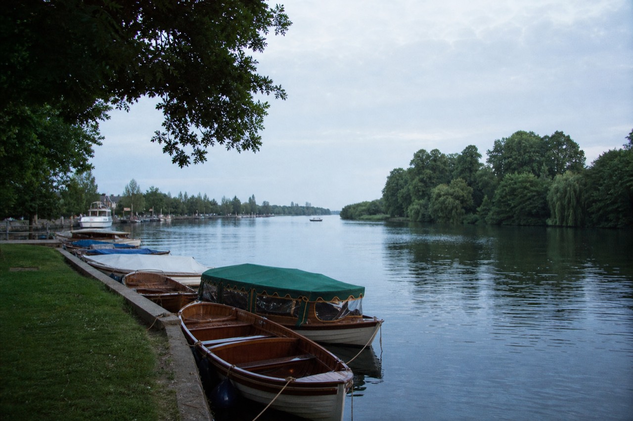A wide straight reach of the upper Thames between wooded banks on a still summer morning, wooden launches moored along a grass bank