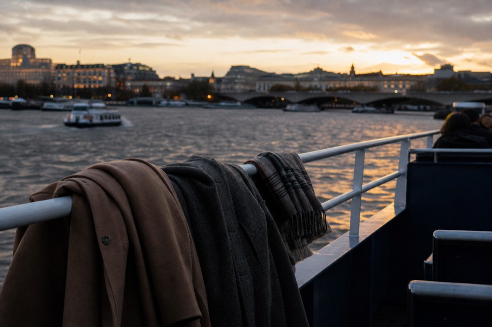 Coats and a folded scarf draped over a boat deck rail at dusk, the river dark below and city lights beginning to show