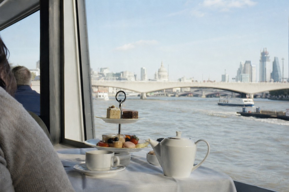 A tiered cake stand and teapot on a white cloth beside a bright boat window, the river and a bridge in full daylight outside