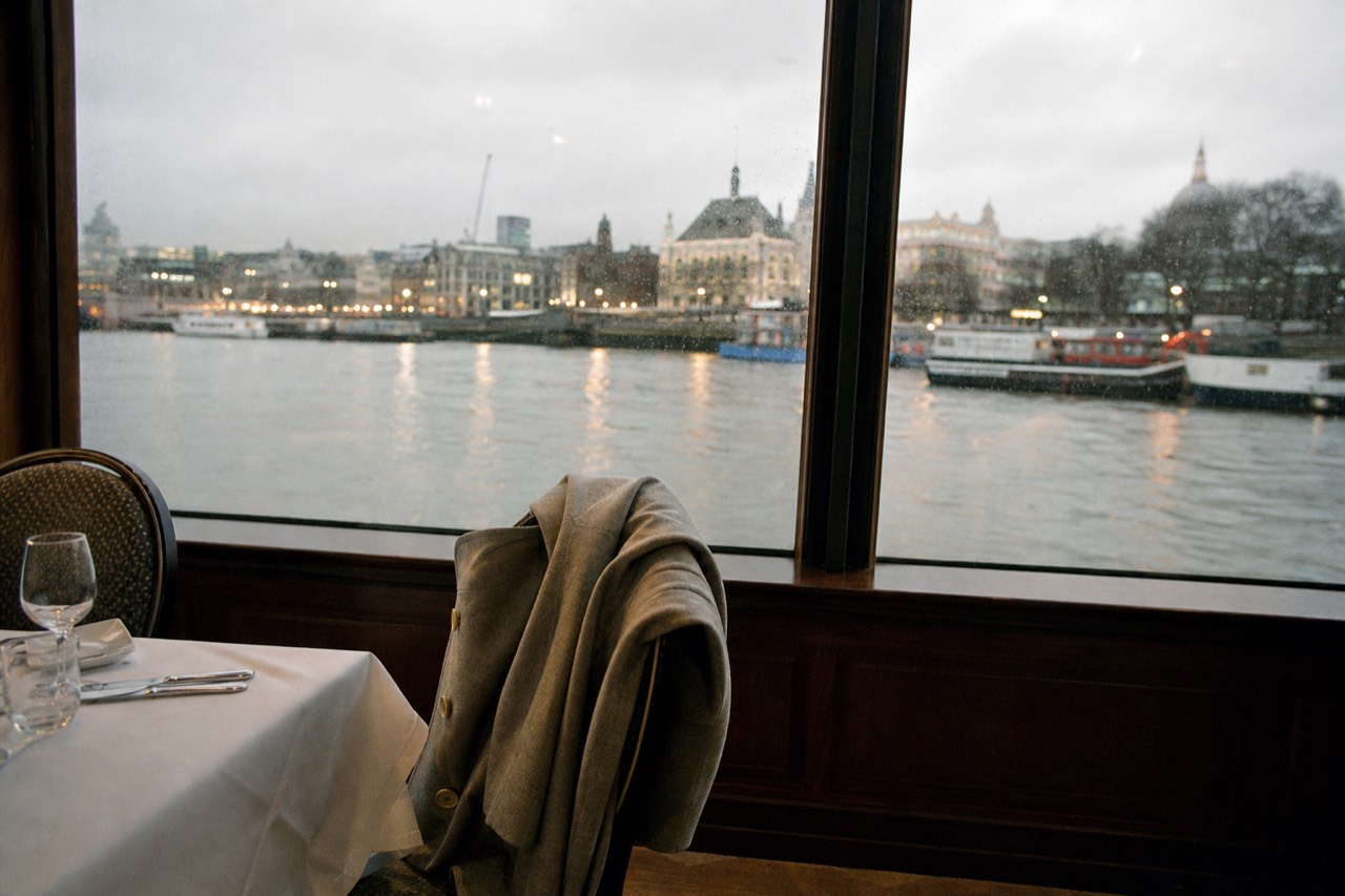 A coat folded over the back of a chair beside a laid dining table on a boat, the dark river and bank lights through the window