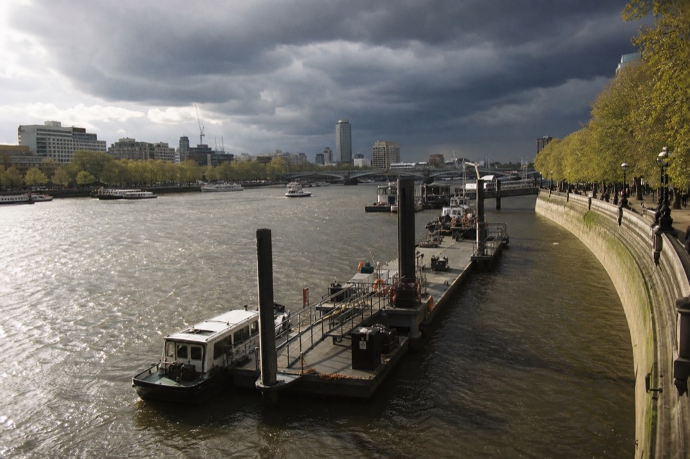 A river pier and its pontoon seen from a bridge above, boats moored alongside and the embankment wall curving away