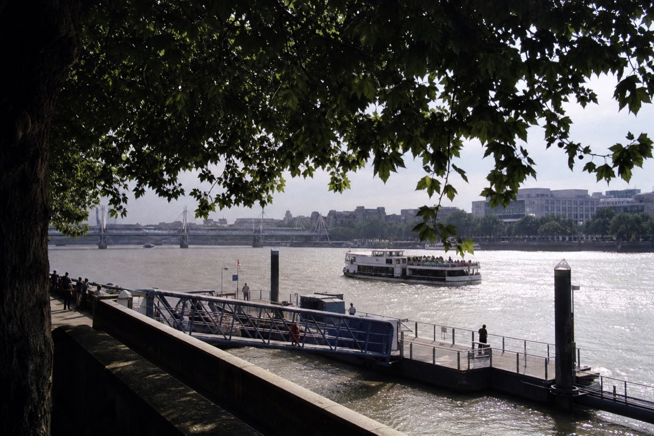 A floating pier pontoon on the north bank of the river with a gangway down and a sightseeing boat approaching in flat afternoon light