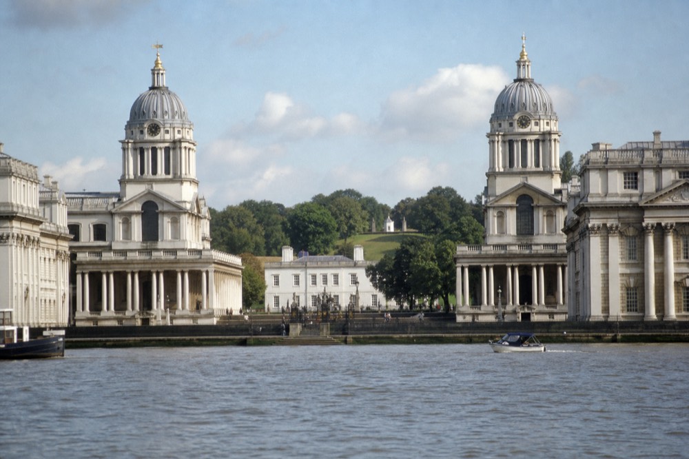 The colonnaded riverfront at Greenwich seen from mid-river on a clear morning, the twin domed blocks framing a smaller white house with green parkland rising behind