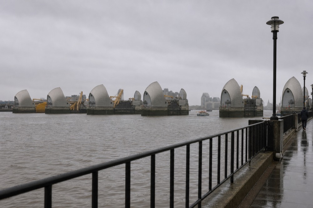 The curved steel shells of the Thames Barrier piers rising from grey water on an overcast afternoon, low industrial banks stretching away either side