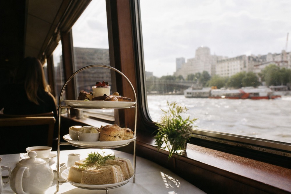 A three-tier cake stand with finger sandwiches, scones and pastries on a laid table beside a boat window, soft afternoon light on the cloth