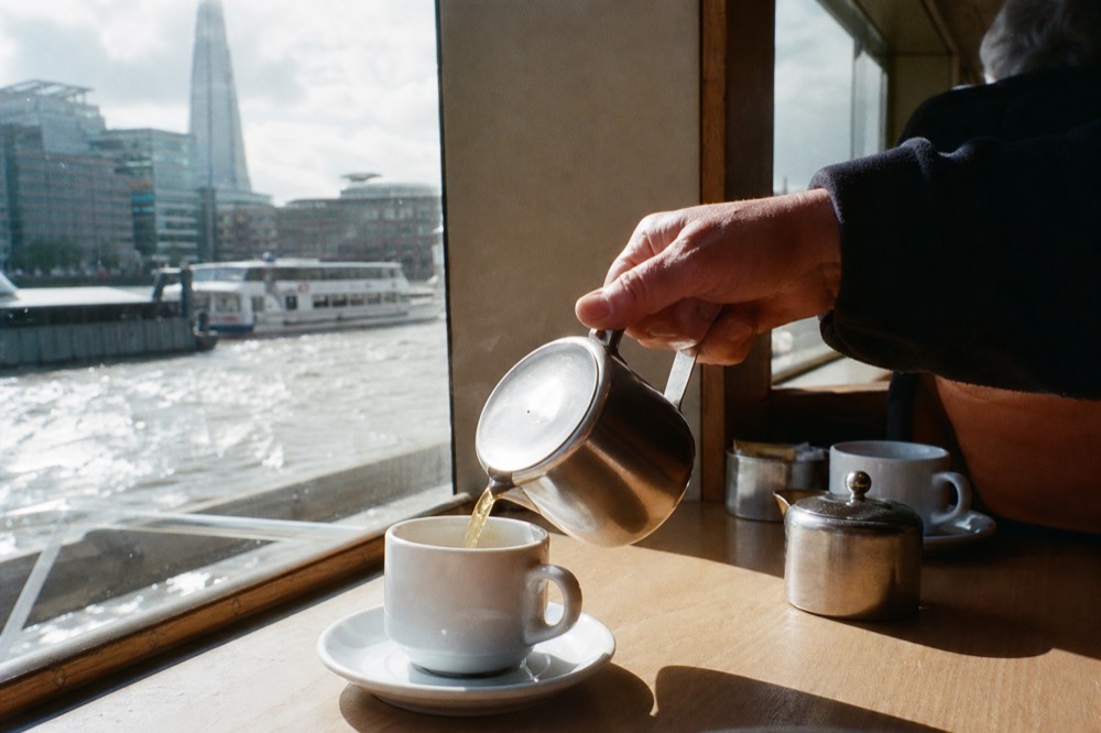 Tea being poured from a metal pot into a white cup on a boat table, river light coming through the window behind