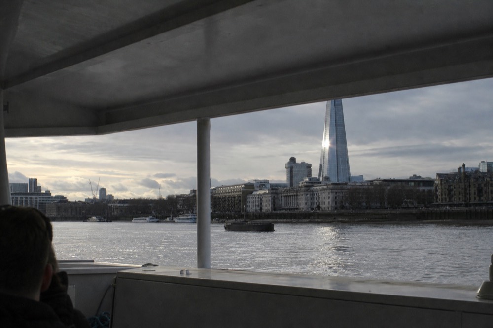 A tall glass tower rising above the south bank of a wide river seen from a boat deck, low afternoon sun catching the glass