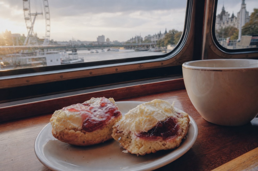 Two halved scones with clotted cream and strawberry jam on a small white plate, a teacup and a boat window edge behind