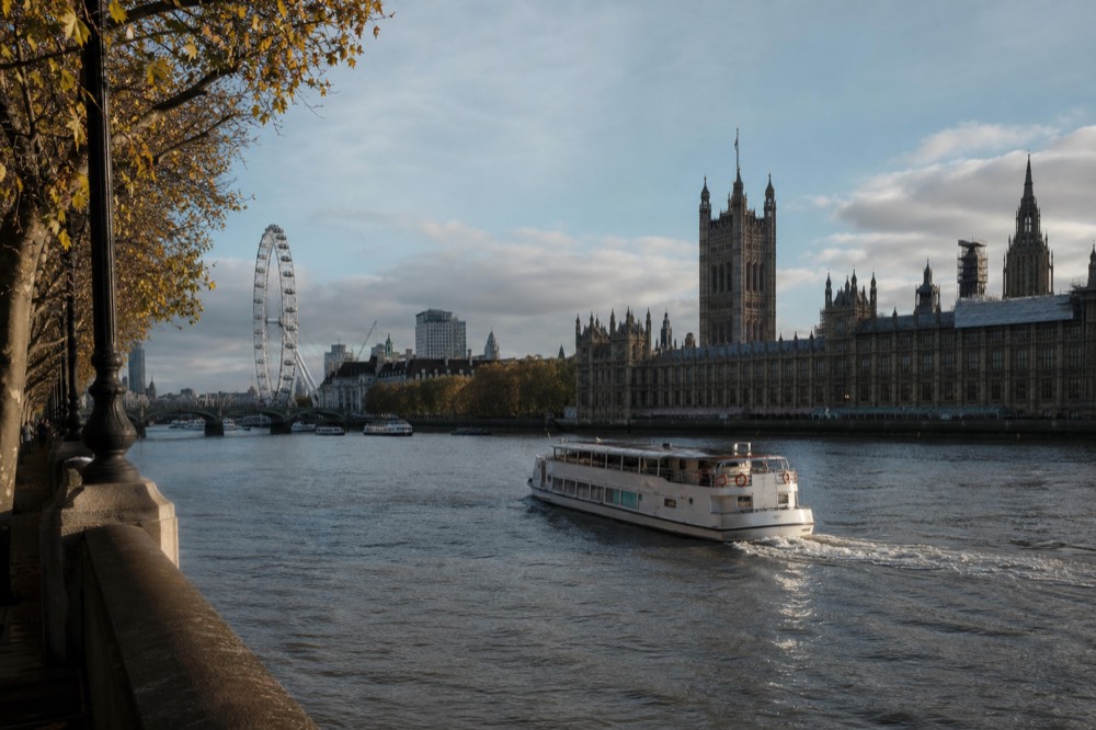 A river boat turning in mid-stream near a Gothic parliament building and a large observation wheel, low afternoon light on the water