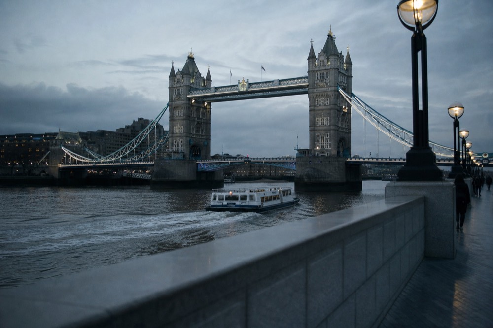 A small sightseeing boat turning in the river just downstream of Tower Bridge, wake curling behind it under grey cloud