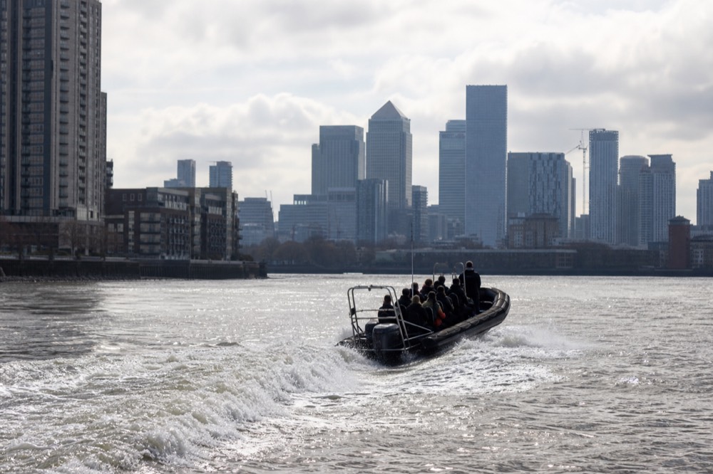 A twelve-seat rigid inflatable boat throwing a broad white wake on the river with the towers of Canary Wharf behind