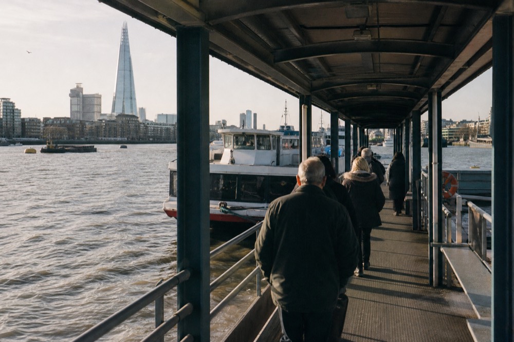 Passengers walking down a covered pier gangway towards a waiting river boat in early morning light beside the embankment