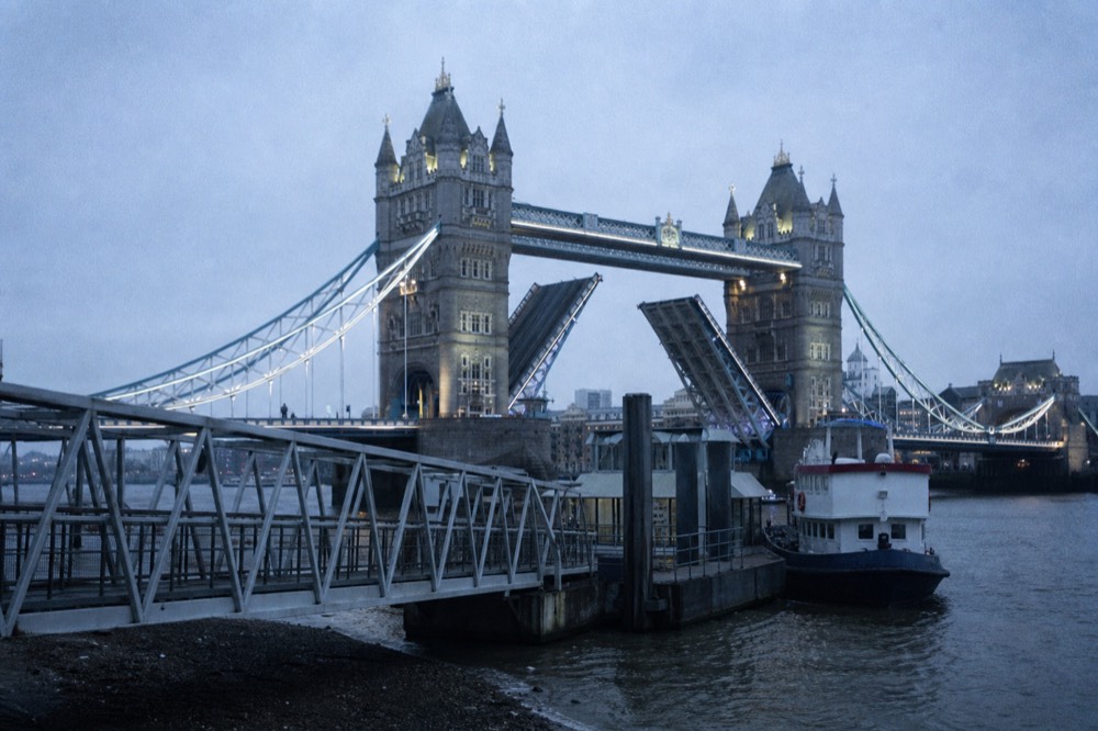 A floating pier pontoon in front of the twin stone towers of a raised bascule bridge, grey water and a moored boat alongside