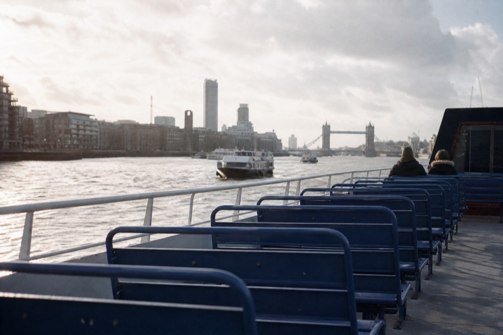 Empty blue bench seating on the open upper deck of a river boat, railings and the wide grey river beyond on a breezy day