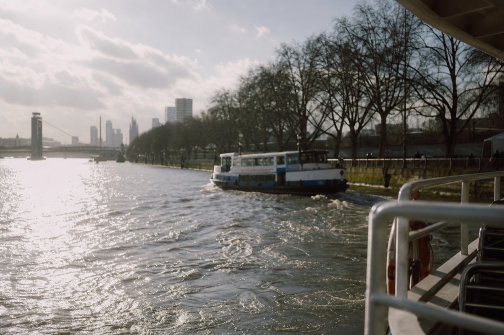 A river boat pulling away from a pier on a clear winter afternoon, low sun flaring off the water and bare trees along the embankment