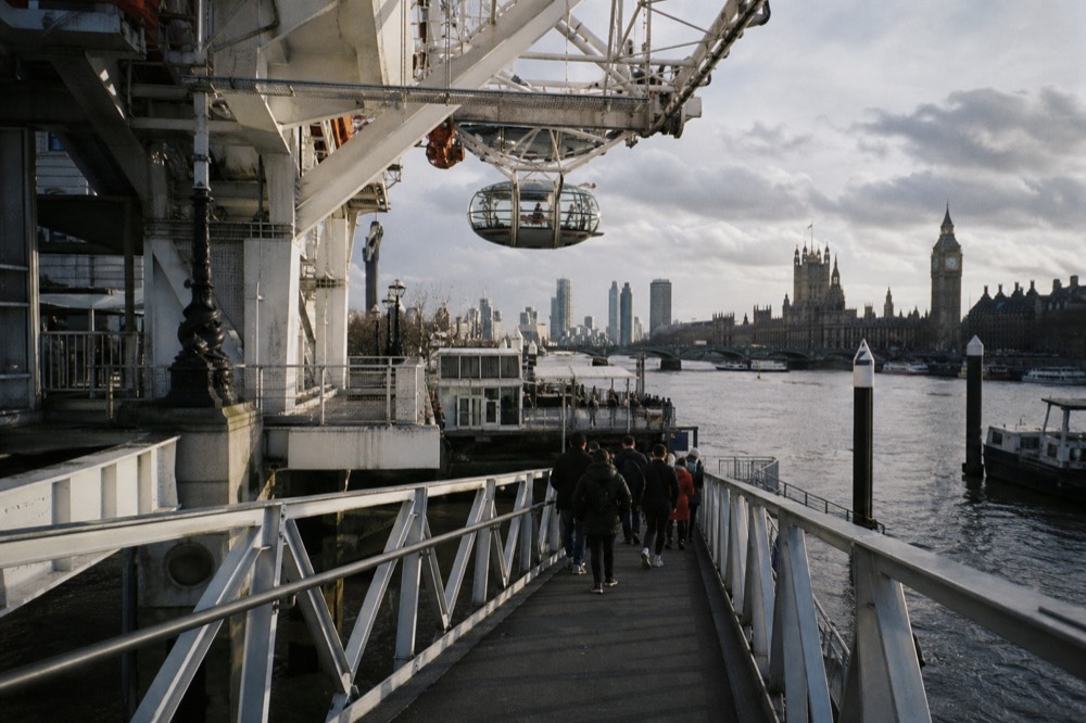 Passengers walking down a metal gangway onto a floating river pier in morning light, the white struts of an observation wheel overhead