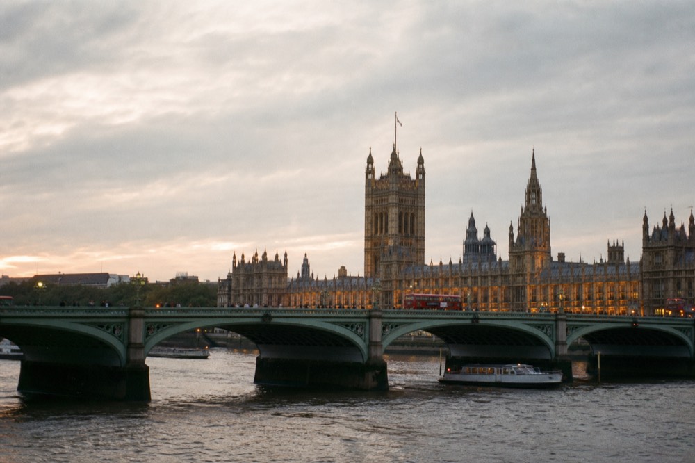 A wide river bridge with pale green ironwork seen low from the water, the pinnacled towers of a parliament building rising behind it