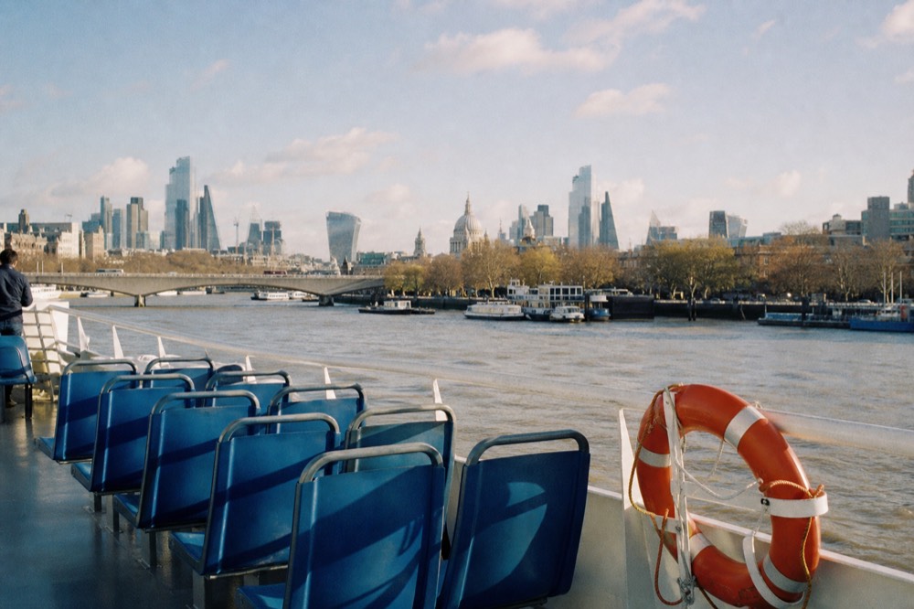 Empty blue bench seats on the open upper deck of a river boat, handrail and life ring in the foreground, city rooftops beyond