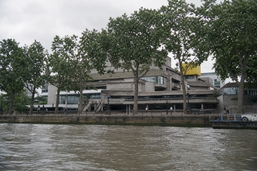 The concrete terraces and walkways of a riverside arts centre seen from mid-river under flat grey cloud, plane trees along the embankment