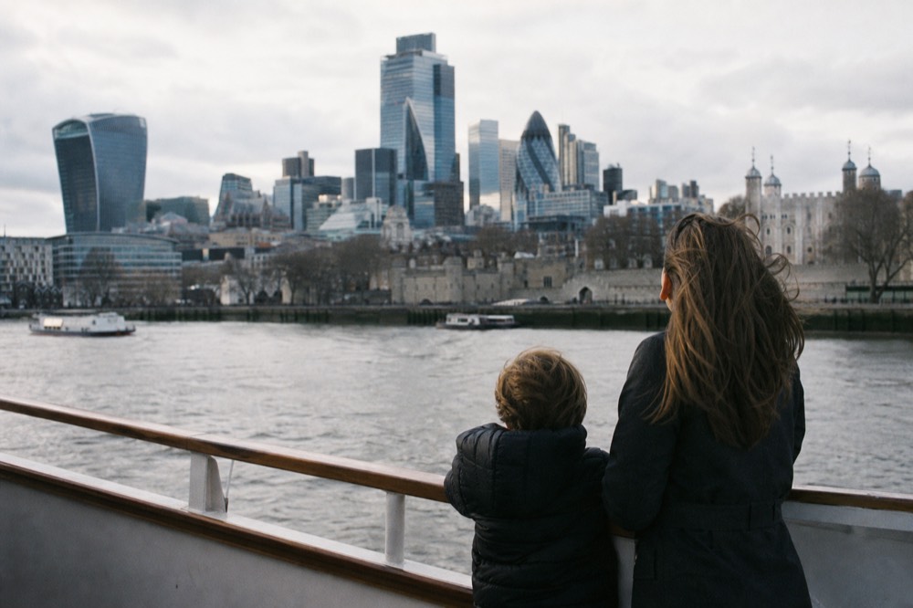 Two small figures at a boat rail from behind, looking out across grey water towards a riverside skyline on an overcast day