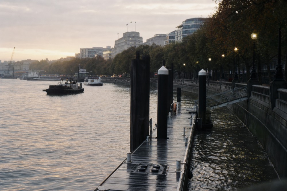 A floating pier pontoon at dusk with wet decking and mooring cleats, the lit south bank walkway curving away behind
