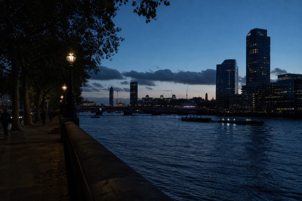The river at blue hour looking downstream, deep blue sky above silhouetted buildings with the first window lights showing