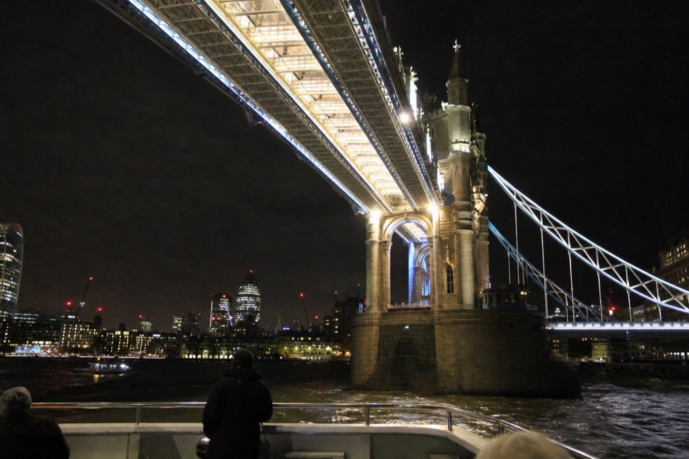 Looking up at the floodlit underside of Tower Bridge from a boat deck at night, ironwork and walkways bright against black sky